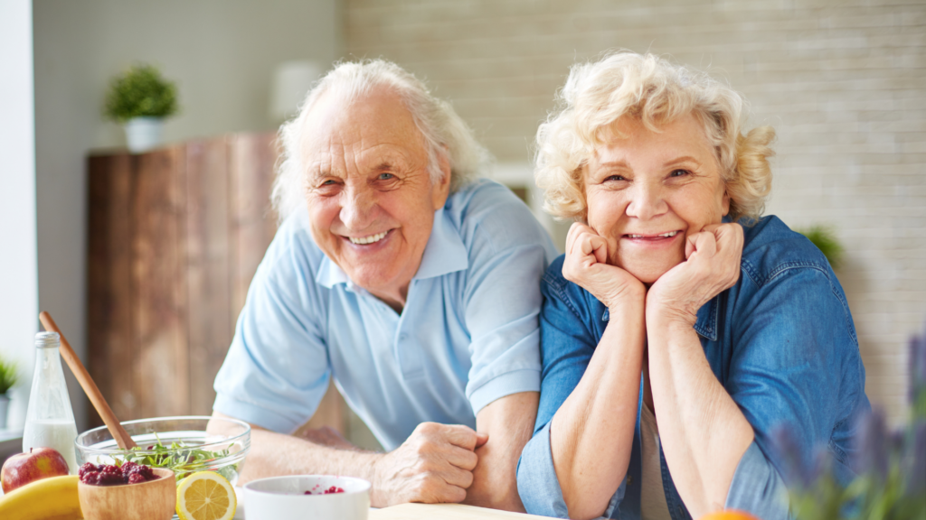 Zu sehen sind zwei Senior/innen, die lächelnd in die Kamera blicken. Vor ihnen befindet sich gesundes Essen.