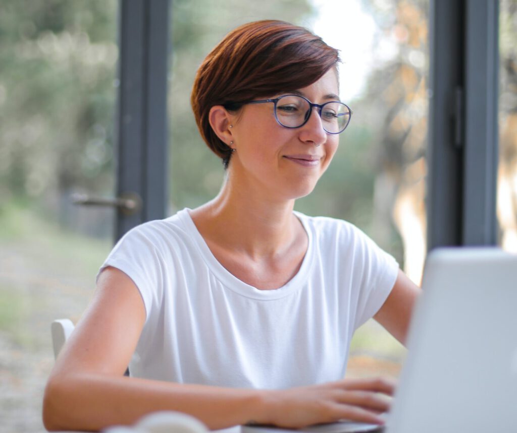 Zu sehen ist eine glückliche junge Dame, die an einem Schreibtisch am Laptop arbeitet.